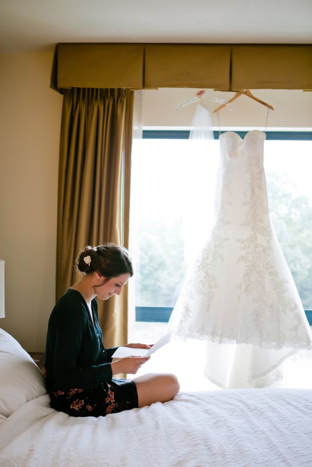 a bride sits and read her love letter from her groom with her wedding dress gracing the window behind her